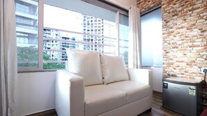 Una habitación con sofá blanco, refrigerador pequeño, pared de ladrillo y ventana con vista a edificios.