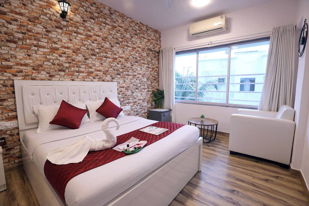 A hotel room with a brick wall, a large bed with red pillows, a white headboard, a small round table, a white sofa, and a window with curtains.