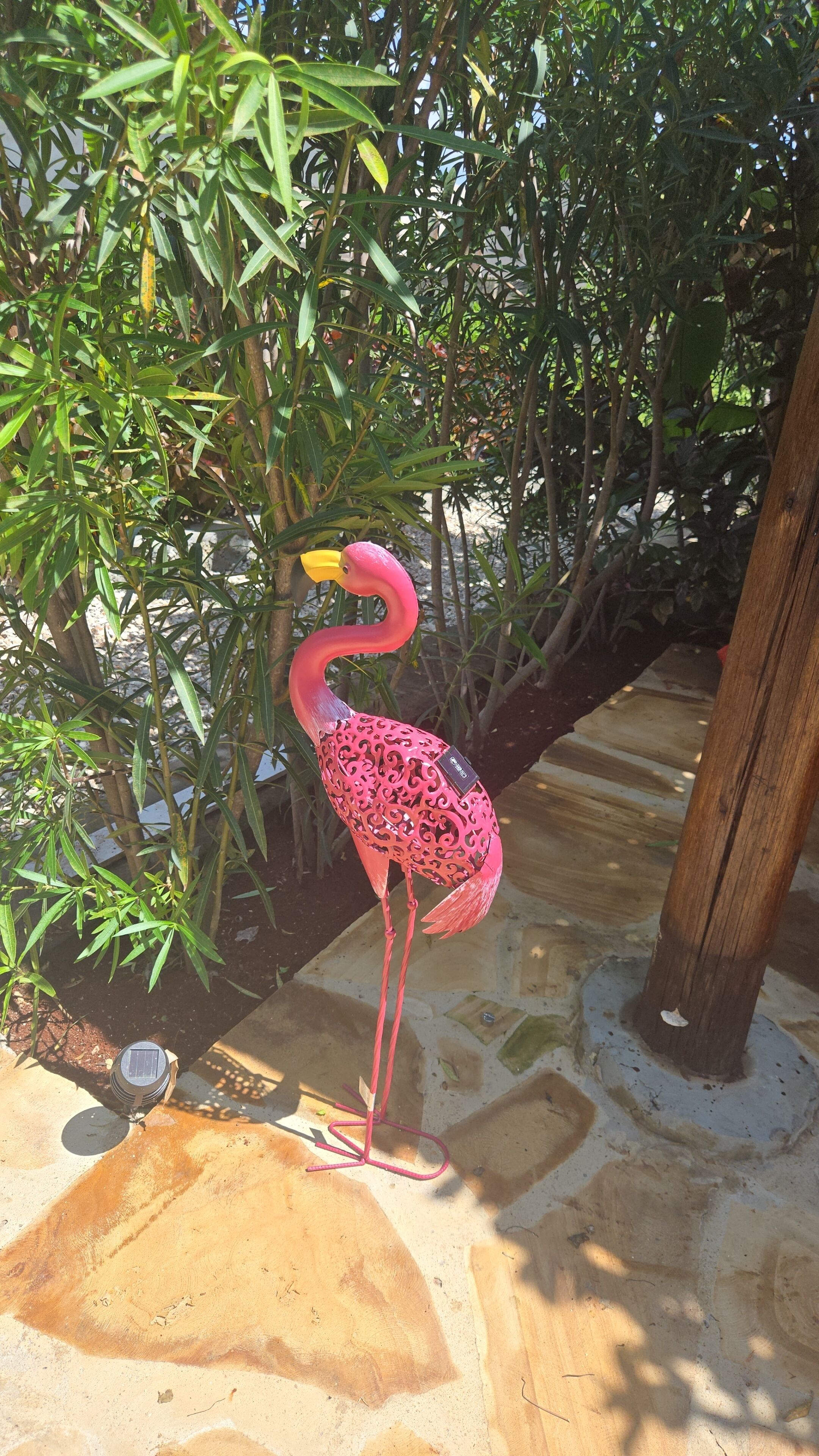 Vaaleanpunainen flamingo-patsas puutarha-alueella, jossa on vihreää kasvillisuutta ja kivinen polku.