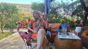 Une personne est assise sur une chaise en osier à l’extérieur, près d’une sculpture de flamant rose. On aperçoit un sol en pierre et une zone abritée recouverte d’une bâche bleue.