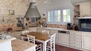 A kitchen with white cabinets, a stone wall, a wooden dining table, and a large window. - Charming house near Chaucre Beach (Saint-Georges-d'Oléron)