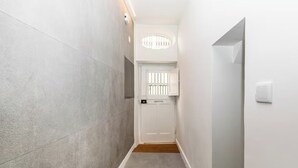 Un couloir étroit, avec une porte blanche au fond, une petite fenêtre et un luminaire au plafond.