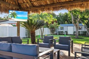 Ein überdachter Außenbereich mit Sitzgelegenheiten, einem Fernsehbildschirm, der eine Strandansicht zeigt, umgeben von Palmen und einem weißen Zaun.