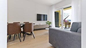 A modern living room with a grey sofa, a dining area with brown chairs, a flat-screen TV mounted on the wall, and a sliding glass door leading to an outdoor area. - Luxury Living at Horizon Retreat - Premium Two Bedroom Apartment (Marina)