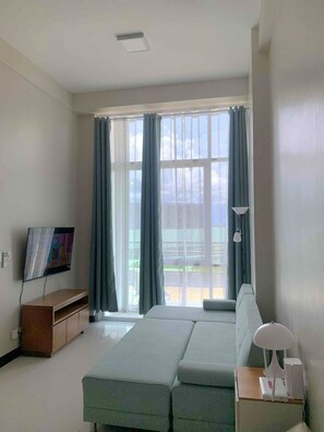 A modern hotel room with a large window, a flat-screen TV, a sofa, a bedside table, and a lamp. - Mactan Newtown 1BR Poolside Stay Near Beach & Airport (Lapu-Lapu)