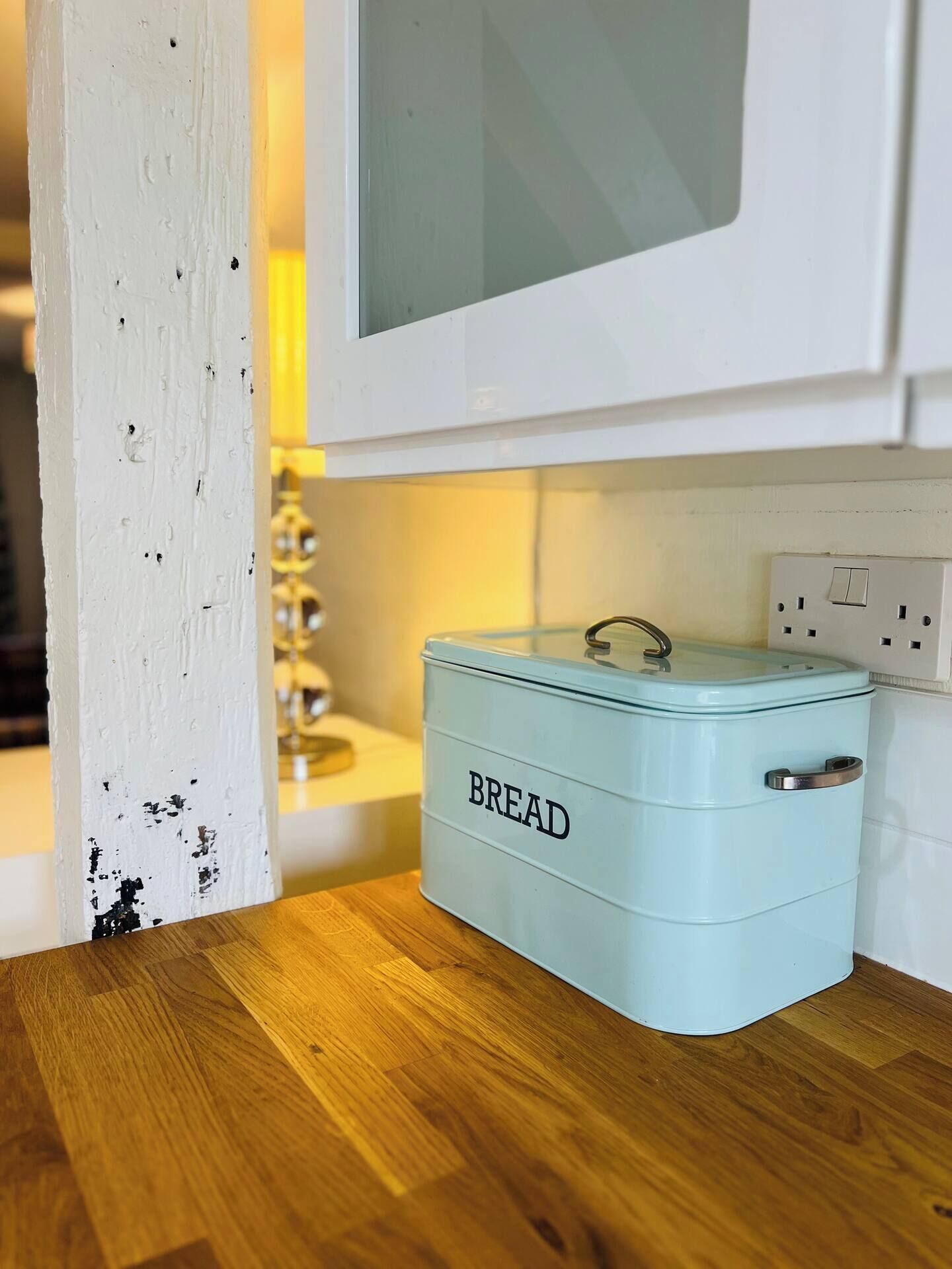 A kitchen with a light blue bread container labeled 'BREAD' on a wooden countertop, a white cabinet above, and a white door with visible wear.