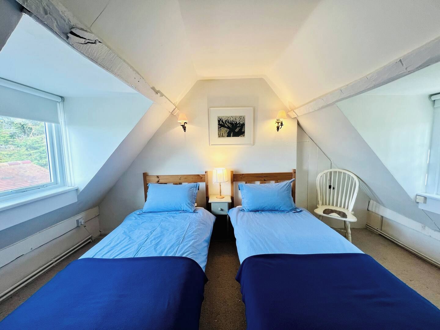 A small, attic-style bedroom with two beds, a white chair, and a painting on the wall.