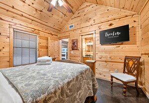 A wooden cabin interior with a bed, a chair, a TV, and a bathroom visible through an open door. - ~2 Acres | Hot Tub | Fire Pit | Near Beavers Bend (Broken Bow)