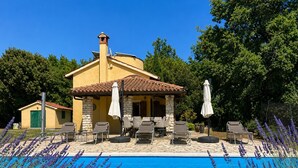 Un área junto a la piscina con sillones de descanso y sombrillas, un edificio de piedra con techo de tejas y una piscina azul cristalina.