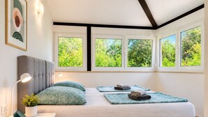 Un dormitorio con cama, lámparas de noche, una mesita de noche, una planta en maceta y una ventana con vista a la vegetación.