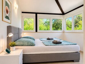 Ein Schlafzimmer mit Bett, Nachttischlampen, einem Nachttisch, einer Topfpflanze und einem Fenster mit Blick auf Grünflächen.