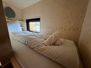 A single bed with white bedding, a window with blinds, and a wooden wall with a leaf decoration. - Tiny Kids Cottage 2+2 (Kaatsheuvel)