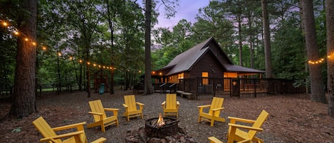 Une cabane entourée d’arbres, avec un foyer et des chaises jaunes au premier plan.
