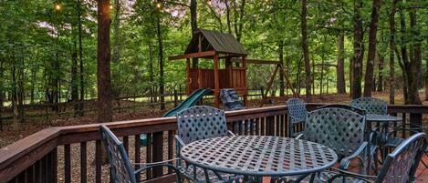A wooden deck with metal chairs and tables, surrounded by a forest.