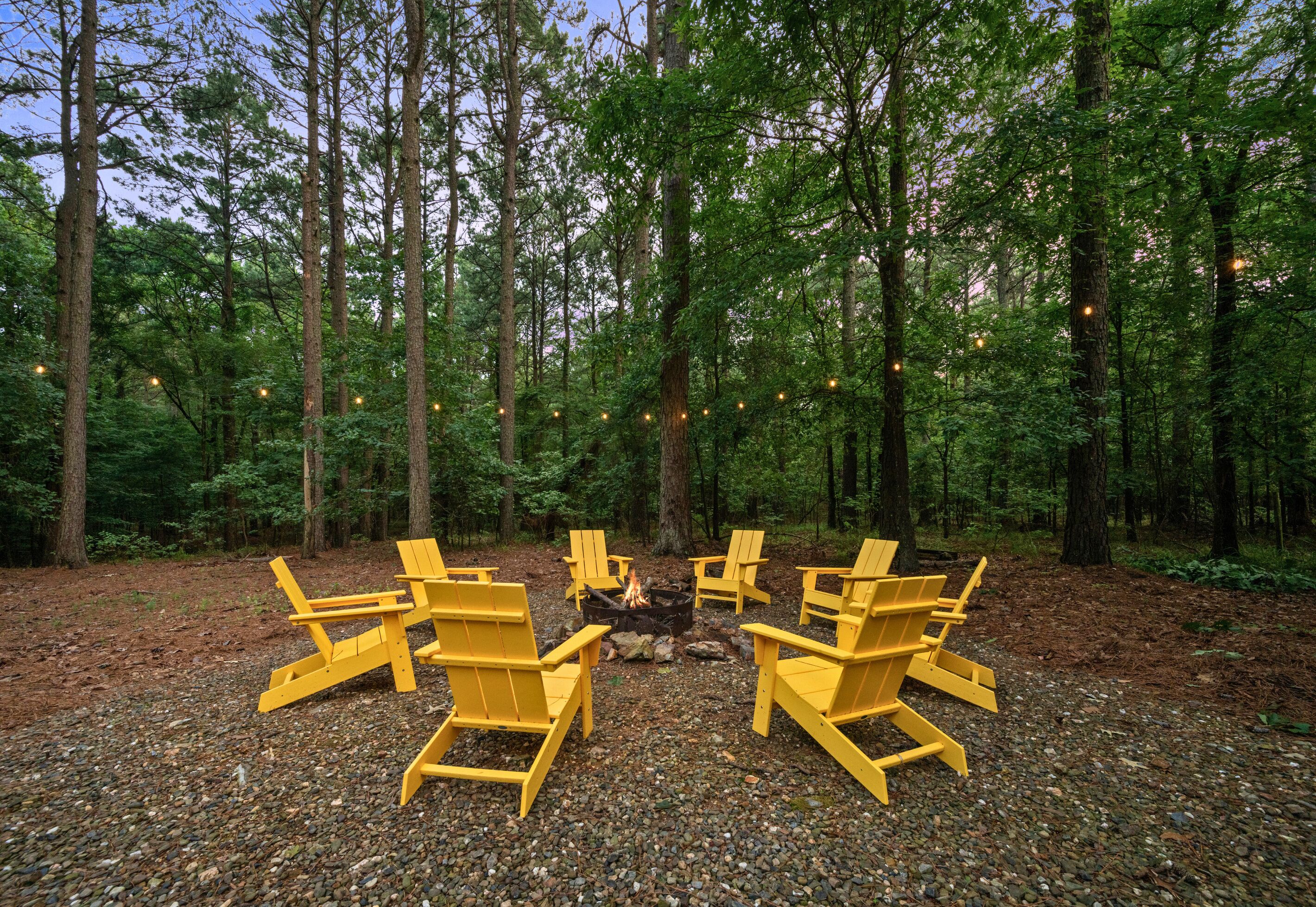 A forest setting with a fire pit and yellow chairs arranged around it.