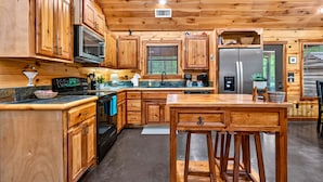 Interior de una cabaña de madera con cocina que incluye armarios de madera, un microondas, estufa, refrigerador y una zona de comedor con taburetes.