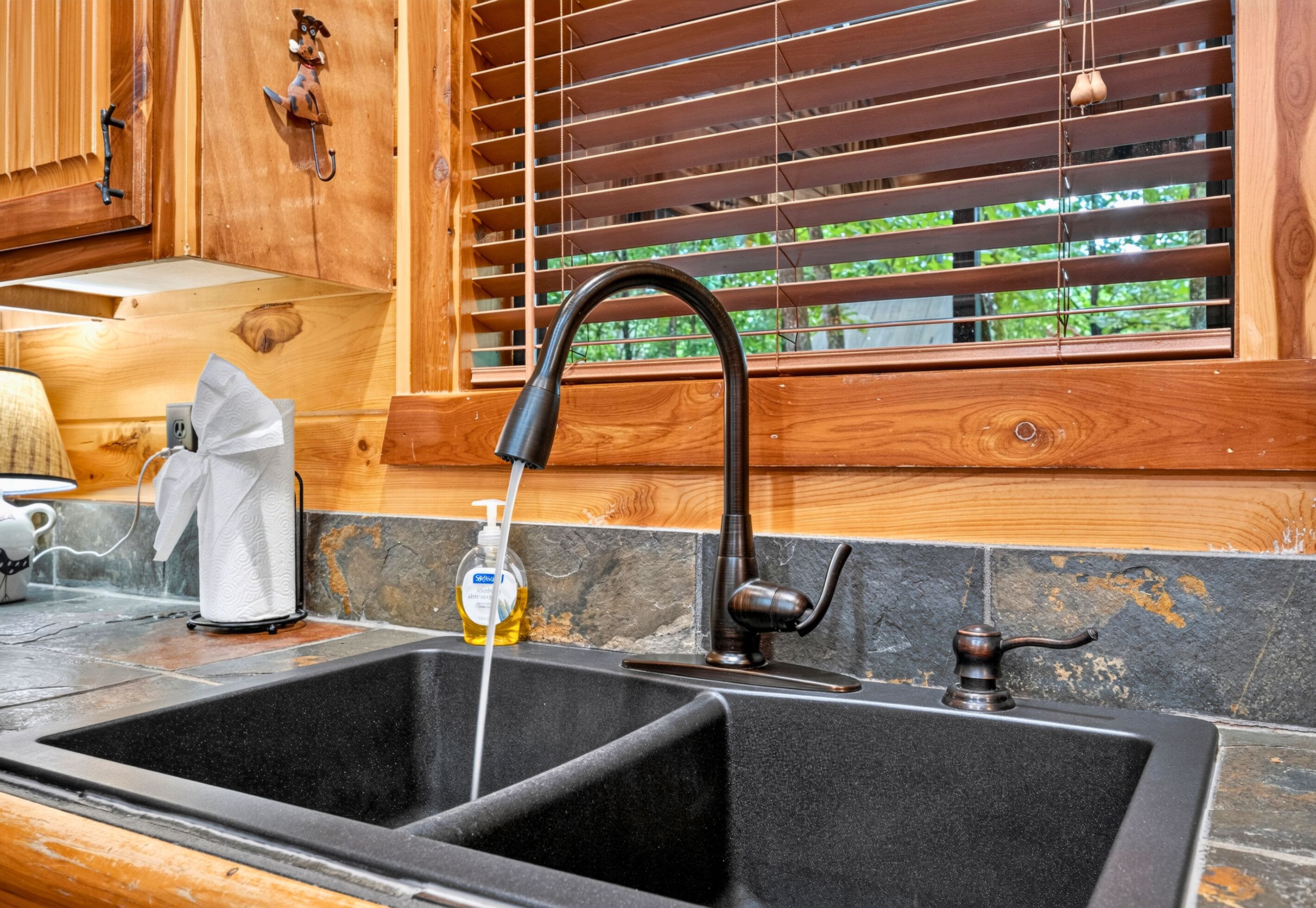 A kitchen with a black sink, wooden cabinets, a window with blinds, and a bottle of dish soap.