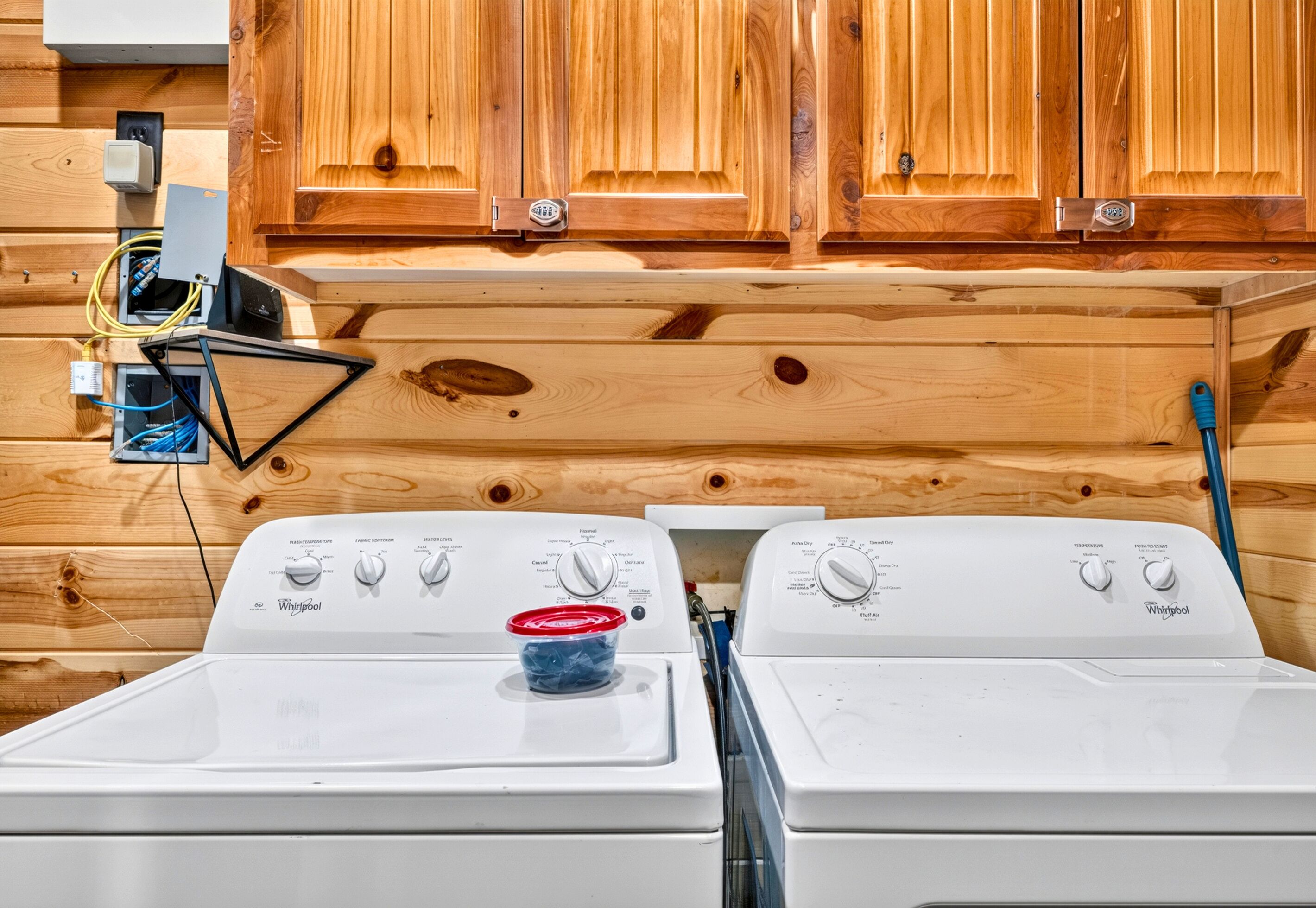 Un área de lavandería con dos lavadoras Whirlpool blancas, armarios de madera arriba y un enchufe conectado.