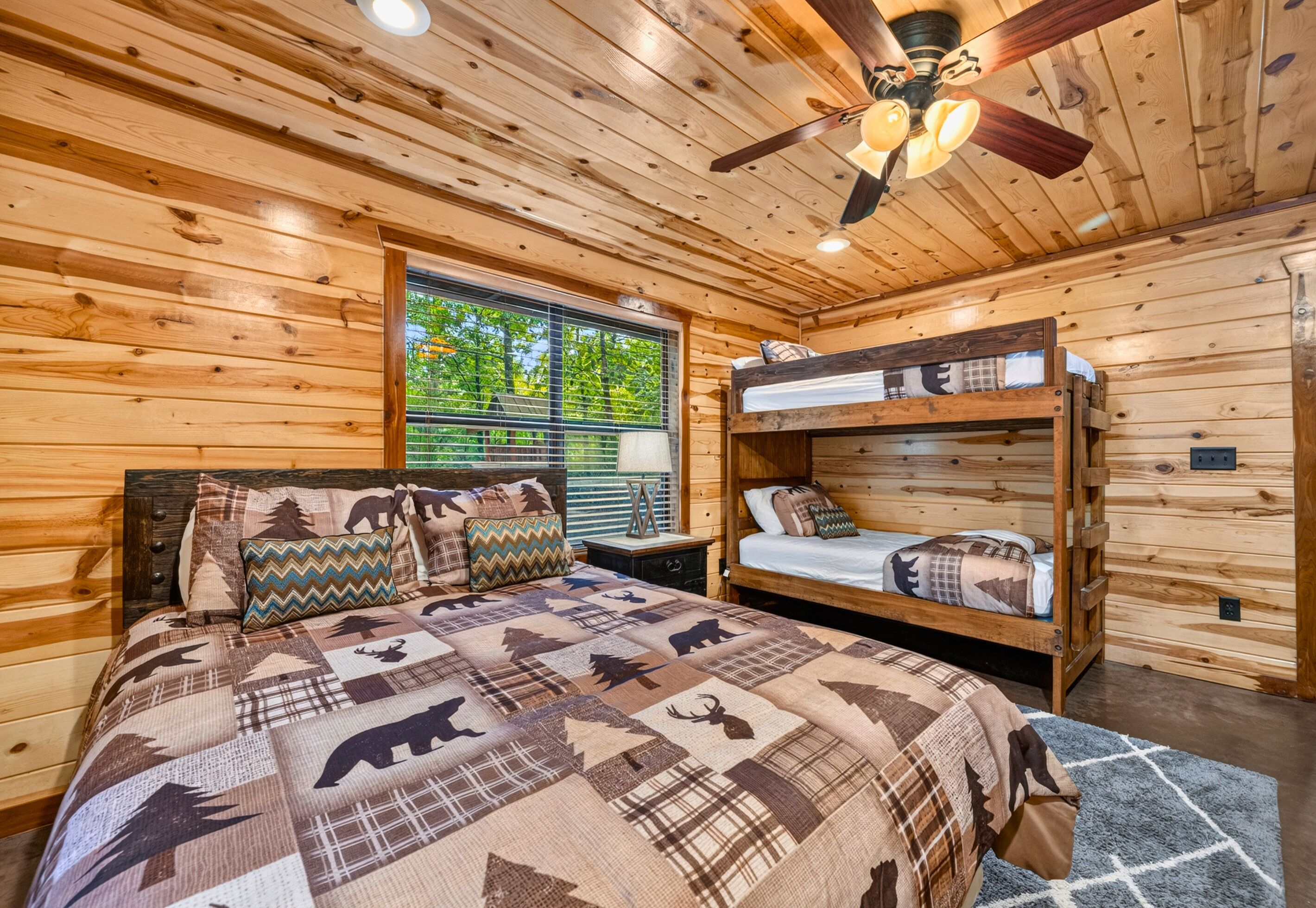 A wooden cabin bedroom with a bunk bed, a large bed with a patterned quilt, a ceiling fan, and a window with blinds.