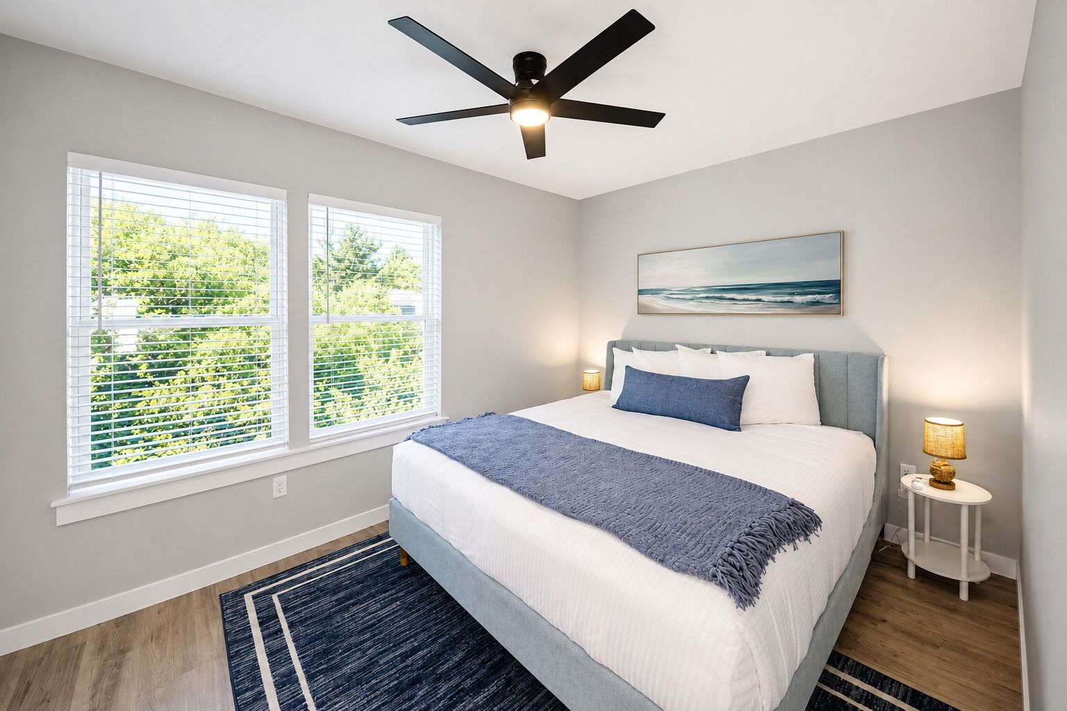 A bedroom with a bed, a ceiling fan, two bedside lamps, a window with blinds, and a painting on the wall.