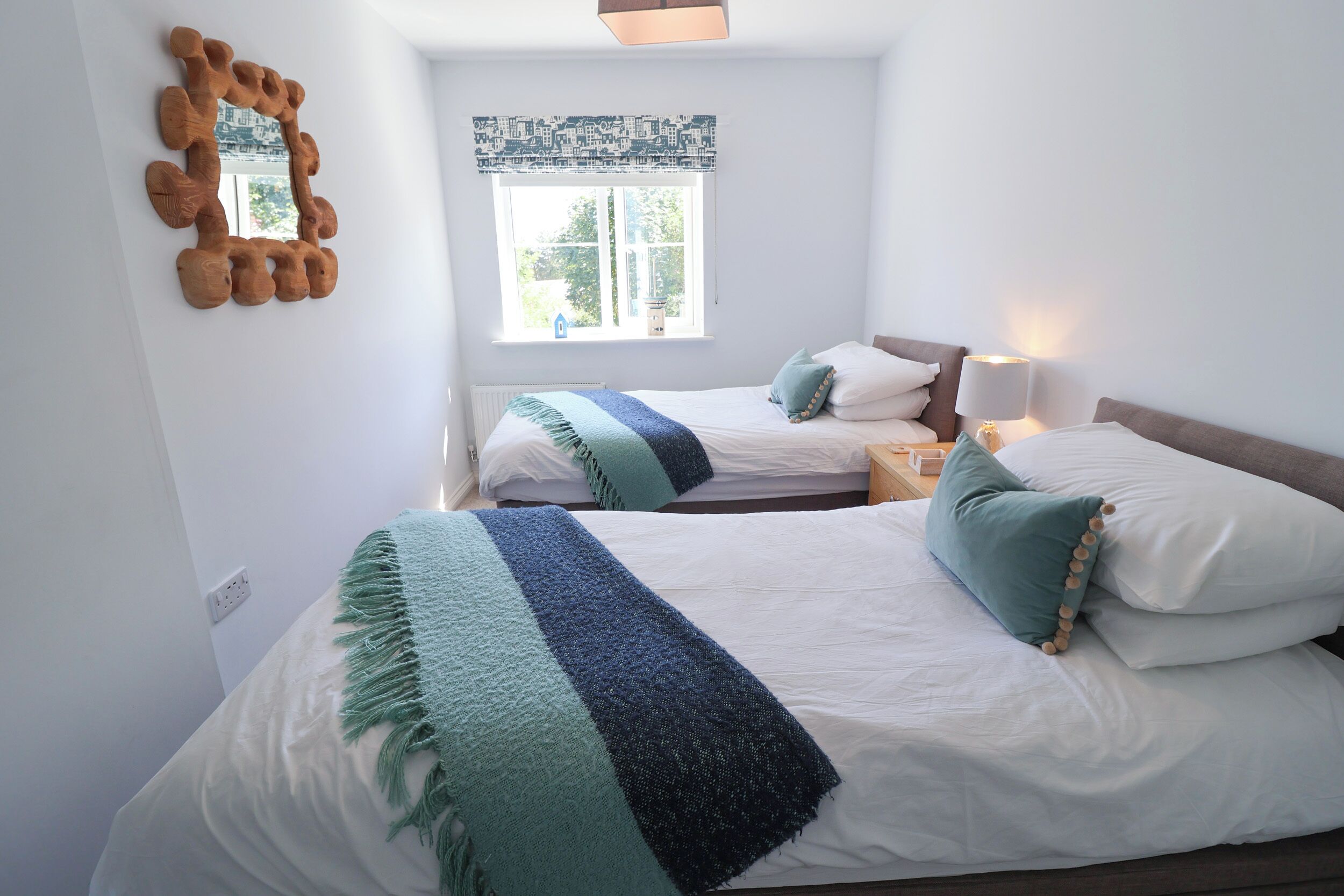 A bedroom with two beds, a window with patterned curtains, a mirror on the wall, and a bedside lamp.