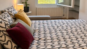 Ein Schlafzimmer mit einem Bett, einem Nachttisch mit Lampe, einer Topfpflanze, einem Fenster mit Blick ins Grüne und einem Kleiderschrank.