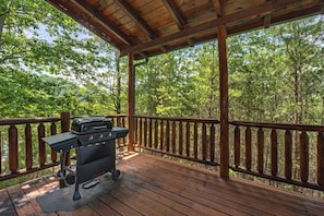 Eine überdachte Terrasse im Freien mit Grill, umgeben von einem hölzernen Geländer und dichtem Wald.