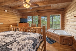 Ein Schlafzimmer in einer Blockhütte mit einem Bett, einem Fernseher, einem Holztisch, einem Stuhl, einem Fenster und einem Badezimmer mit einer Badewanne.