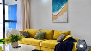 Ett modernt vardagsrum med en gul soffa, en vit matta, en tavla som föreställer en strand och en hängande lampa.