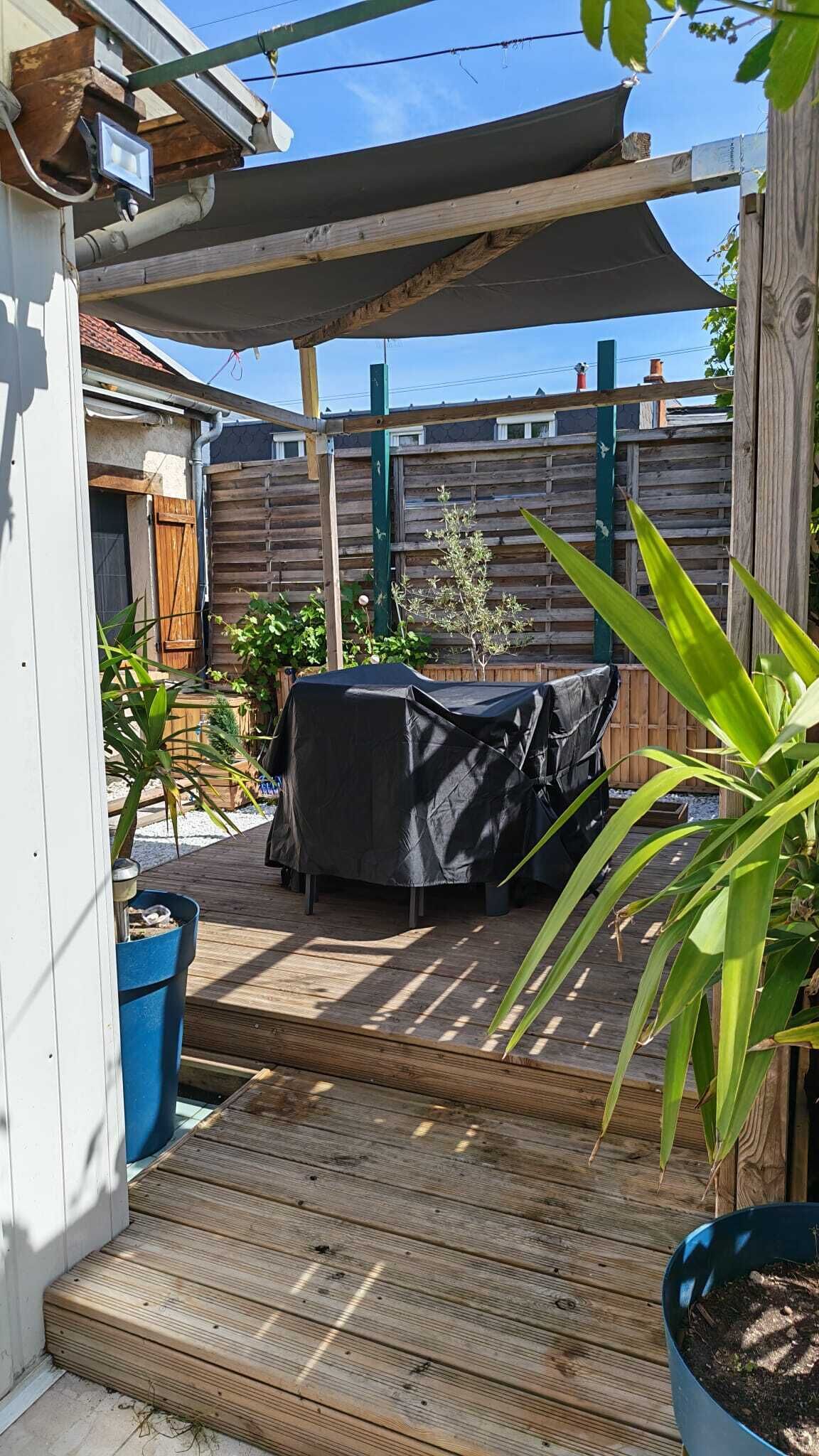 A covered patio with a grill, wooden deck, potted plants, and a wooden fence.