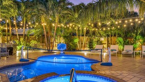Una piscina con aro de baloncesto, rodeada de palmeras y luces colgantes.