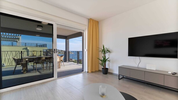 Un soggiorno moderno con una grande TV, un'area pranzo con sedie, una pianta in vaso e un balcone con vista.