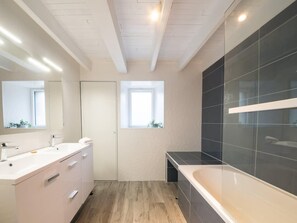 Un baño moderno con un espejo grande, un tocador blanco, una bañera y una ventana.