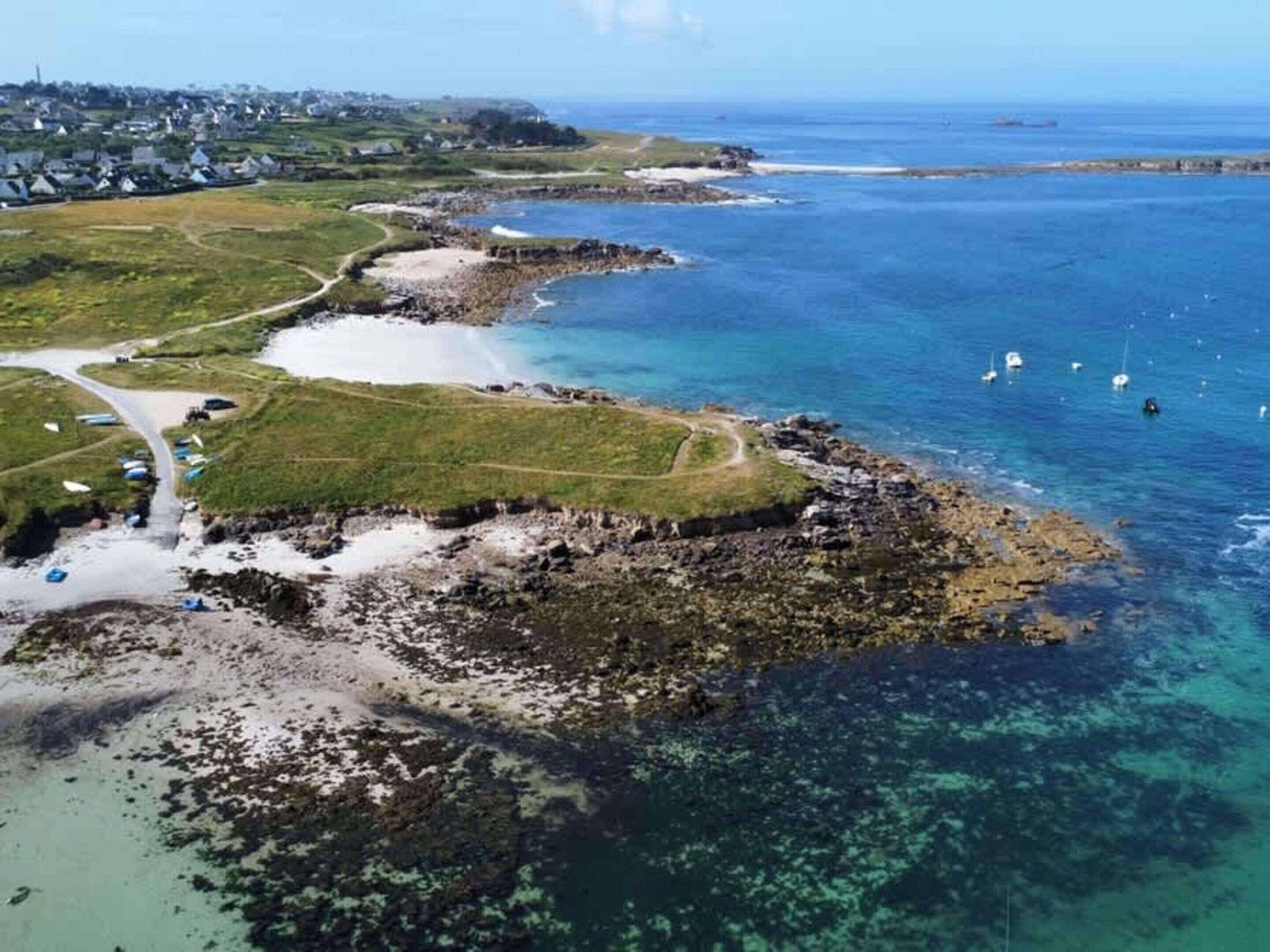 An aerial view of a coastal area with clear waters, boats, and a road leading to a grassy area with buildings in the background.