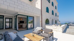 A modern outdoor seating area with a sofa, chairs, and a table, overlooking a building with arched windows and a view of the sea. - Apartment With Pool and hot tub (Konavle)