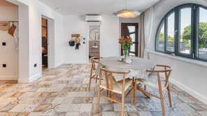 A dining area with a round table, six chairs, a hanging light fixture, and a view through a large window. - Apartment With Pool and hot tub (Konavle)
