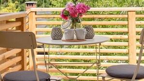 Um deck de madeira com uma mesa e cadeiras, um vaso com flores e montanhas ao fundo.