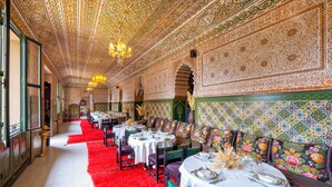 Un amplio comedor con intrincados diseños en el techo, candelabros y una alfombra roja. Las mesas están dispuestas con manteles blancos y cubertería.