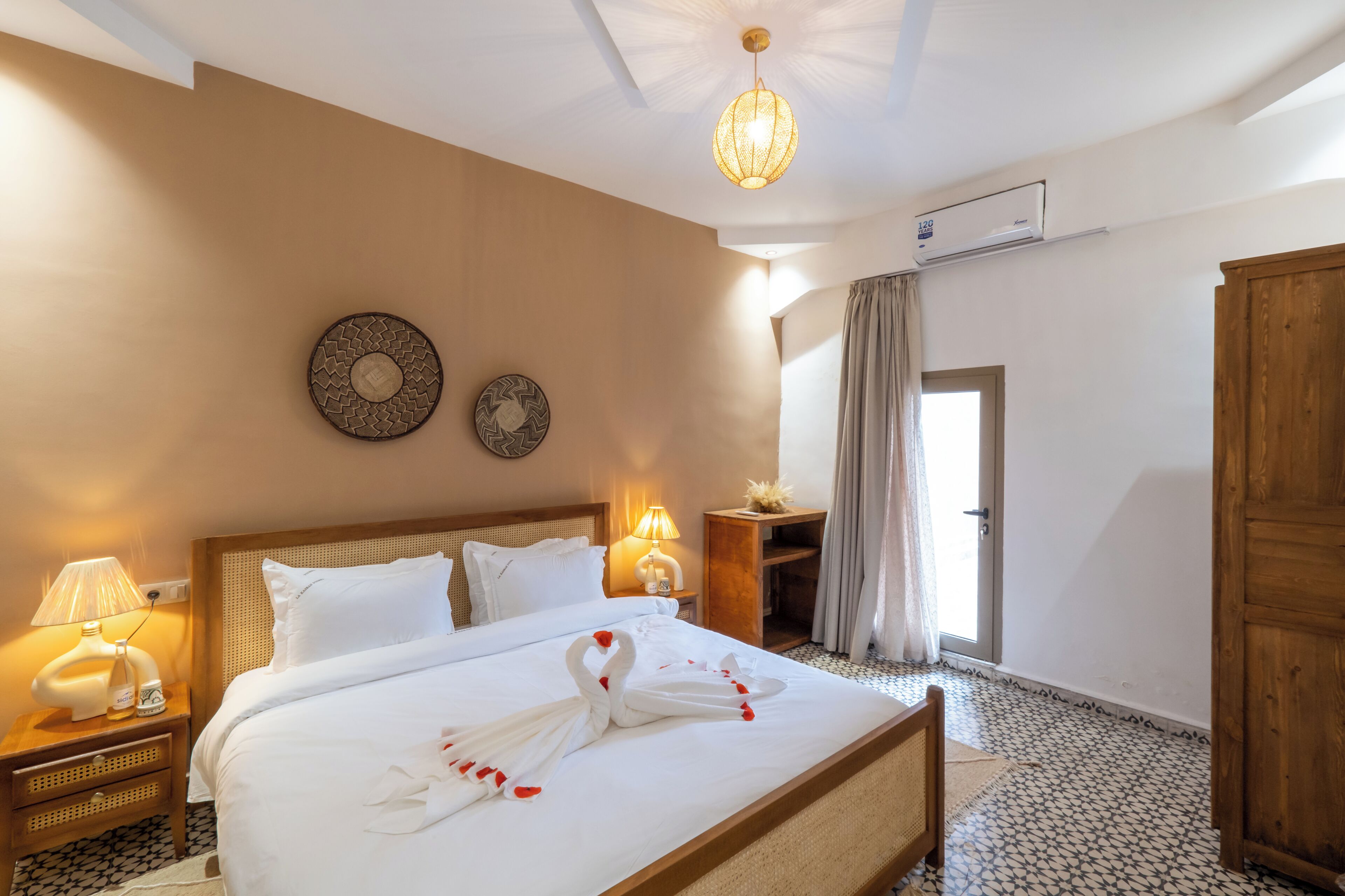 Habitación de hotel con una cama bien hecha, una mesita de noche con lámpara, un reloj de pared y una puerta que da a otra habitación.