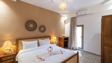 Habitación de hotel con una cama bien hecha, una mesita de noche con lámpara, un reloj de pared y una puerta que da a otra habitación.