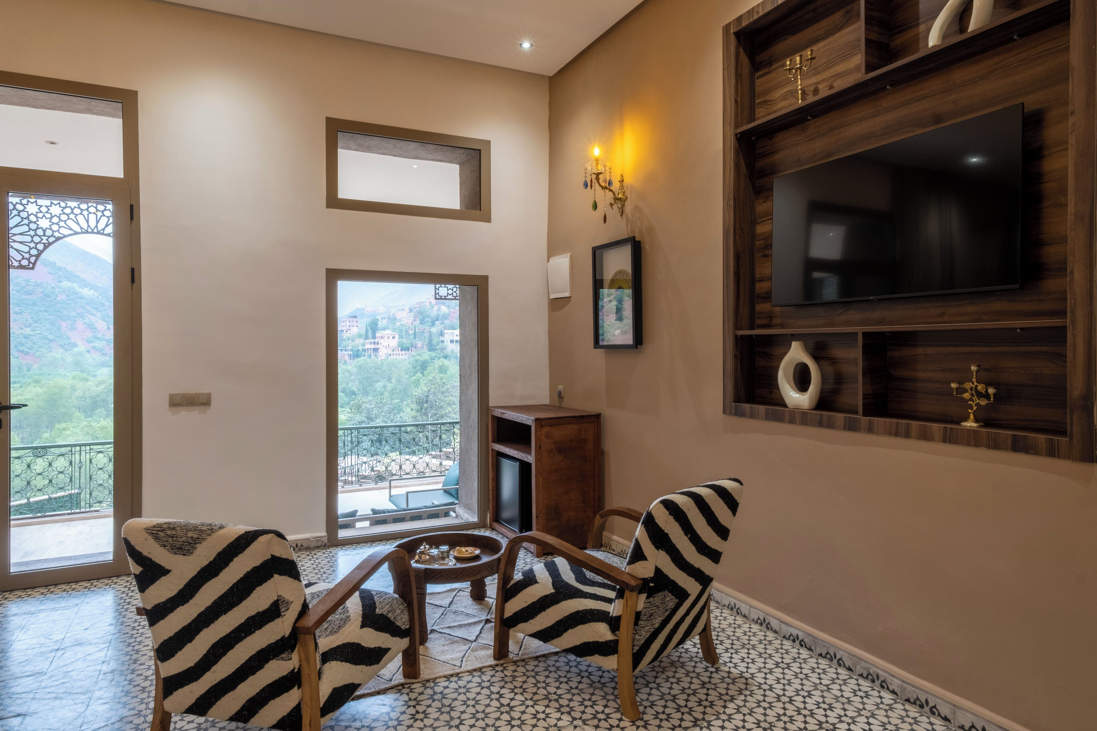 A room with a TV, wooden shelves, a chandelier, and a view of greenery.