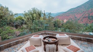 Un balcón con barandilla de vidrio, dos sofás color beige, una pequeña mesa de madera con una bandeja y vistas a un paisaje montañoso.