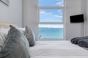 A bedroom with a large window, a bed with striped bedding, and a television on the wall. - North Beach Sea View - Close to Town and Seafront (Tenby)