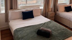 A hotel room with a large bed, a smaller bed, a nightstand, and a view of the city through large windows. - Apartment Sleeps 6 With Air Conditioning (Manchester)