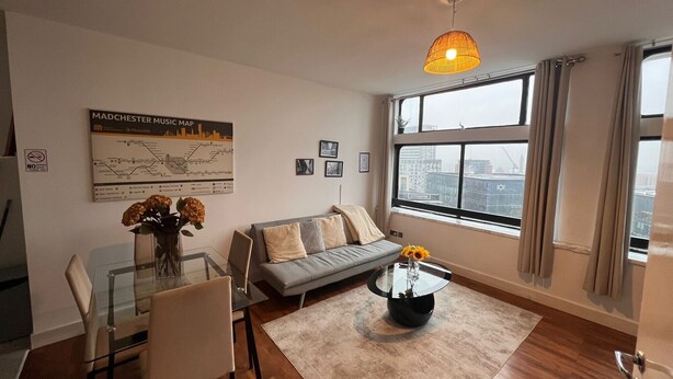 A modern living room with a glass dining table, a grey sofa, a coffee table with flowers, a cityscape view, and a map on the wall. - Apartment Sleeps 6 With Air Conditioning (Manchester)