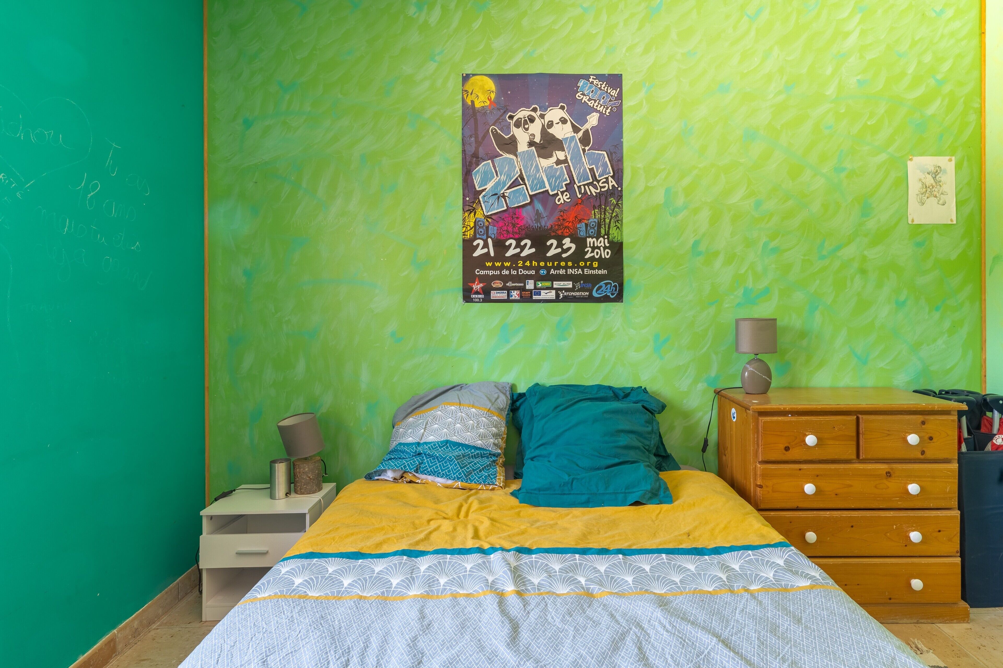Un dormitorio con pared verde, una cama con ropa de cama estampada, un aparador de madera y un cartel en la pared.