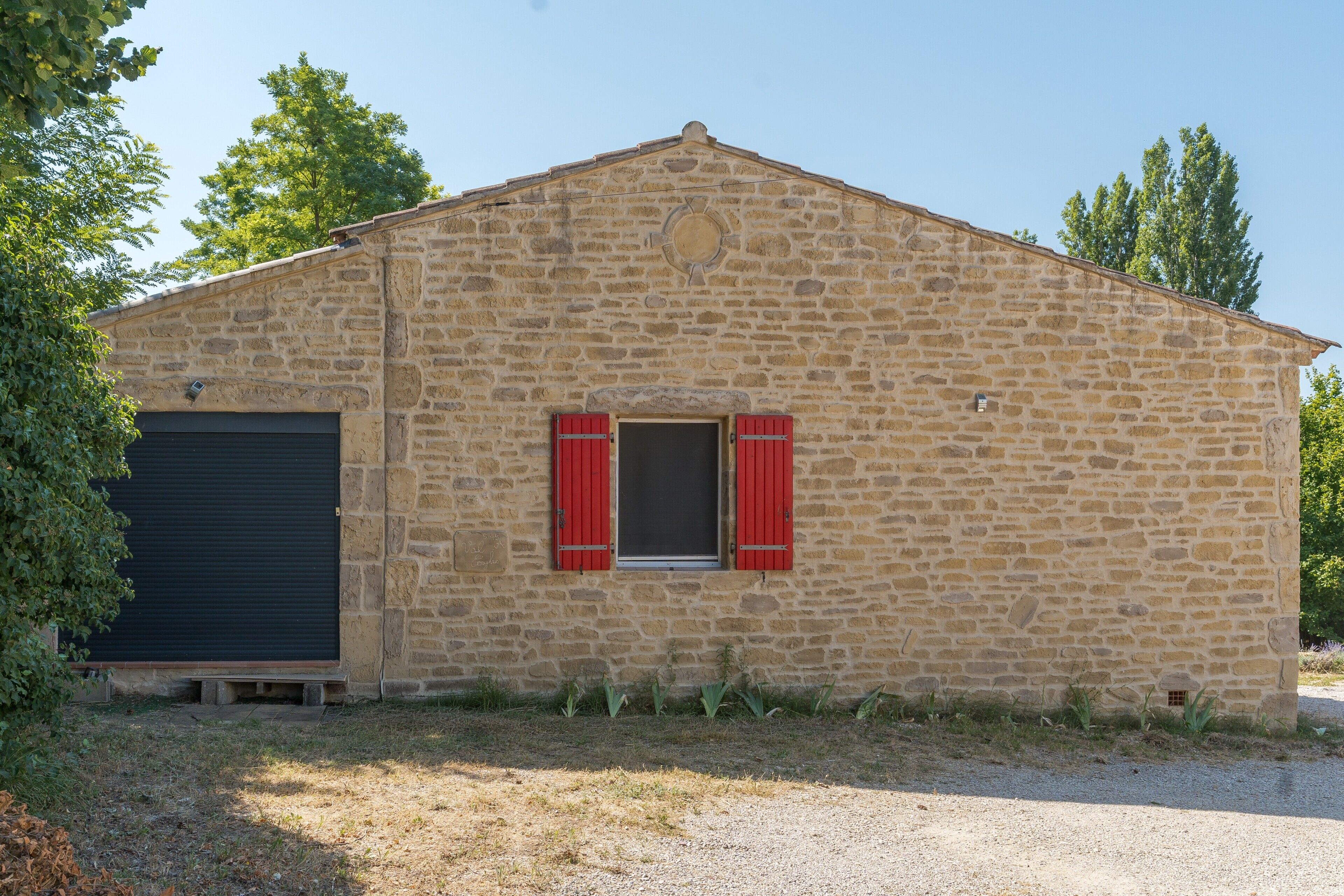 Un bâtiment en pierre avec une porte de garage, une petite fenêtre avec des volets et une allée gravillonnée.