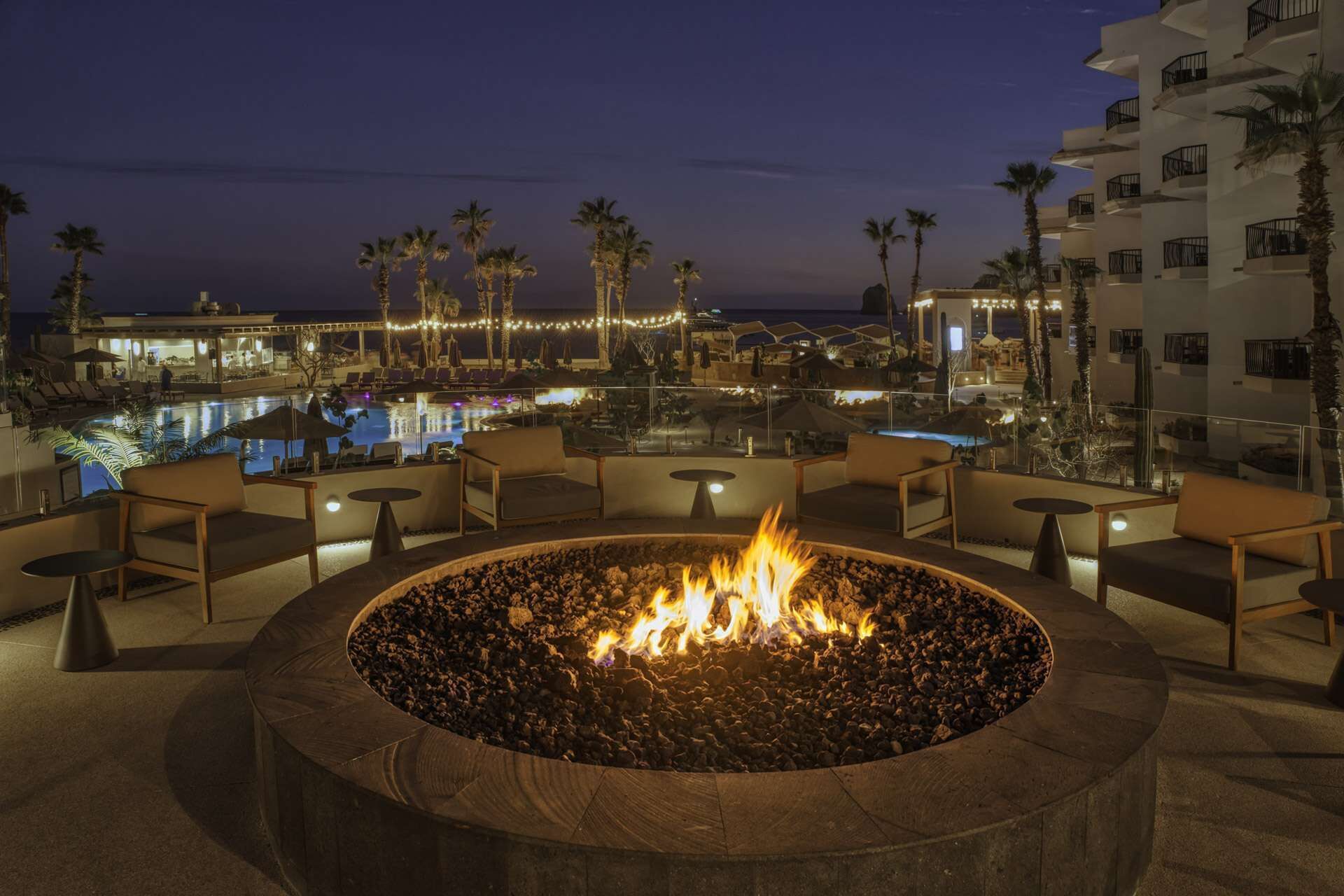 A fire pit surrounded by lounge chairs and tables, overlooking a pool and palm trees.
