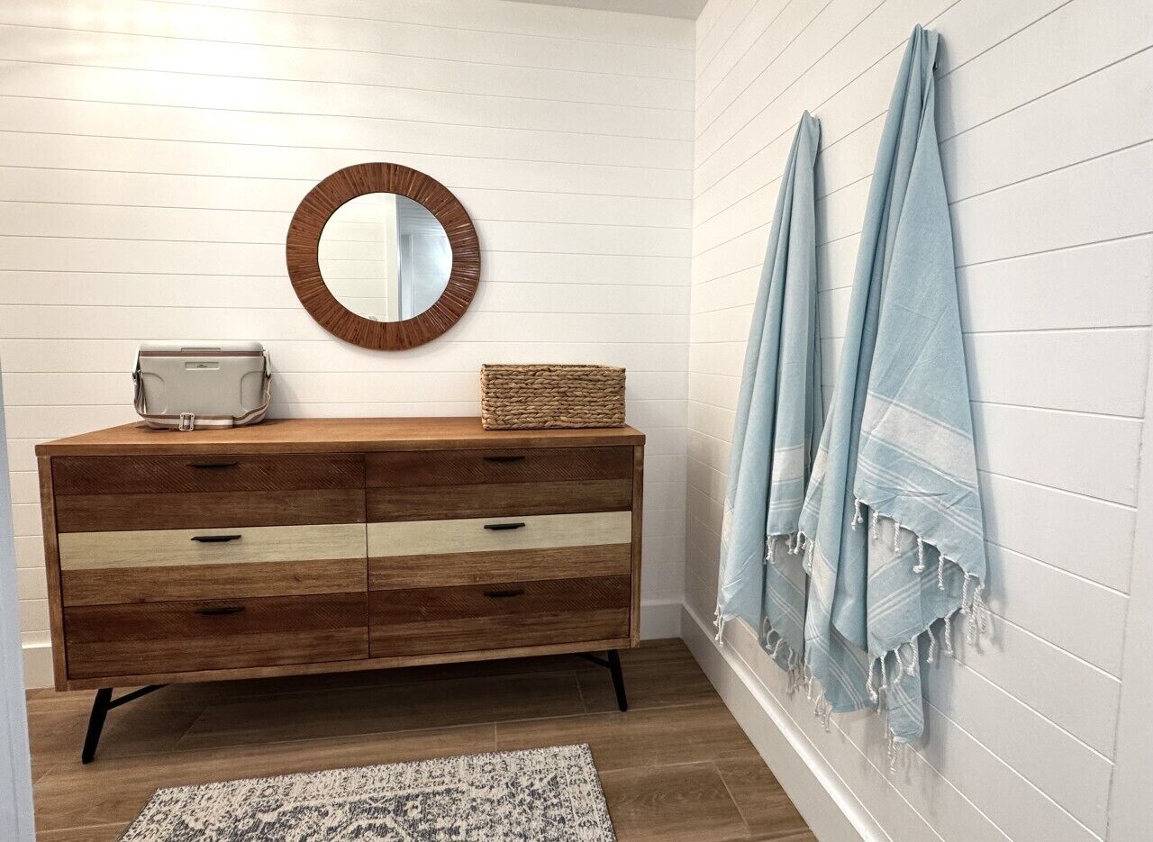 A wooden dresser with drawers, a round mirror, a woven basket, and a blue towel on a wall.