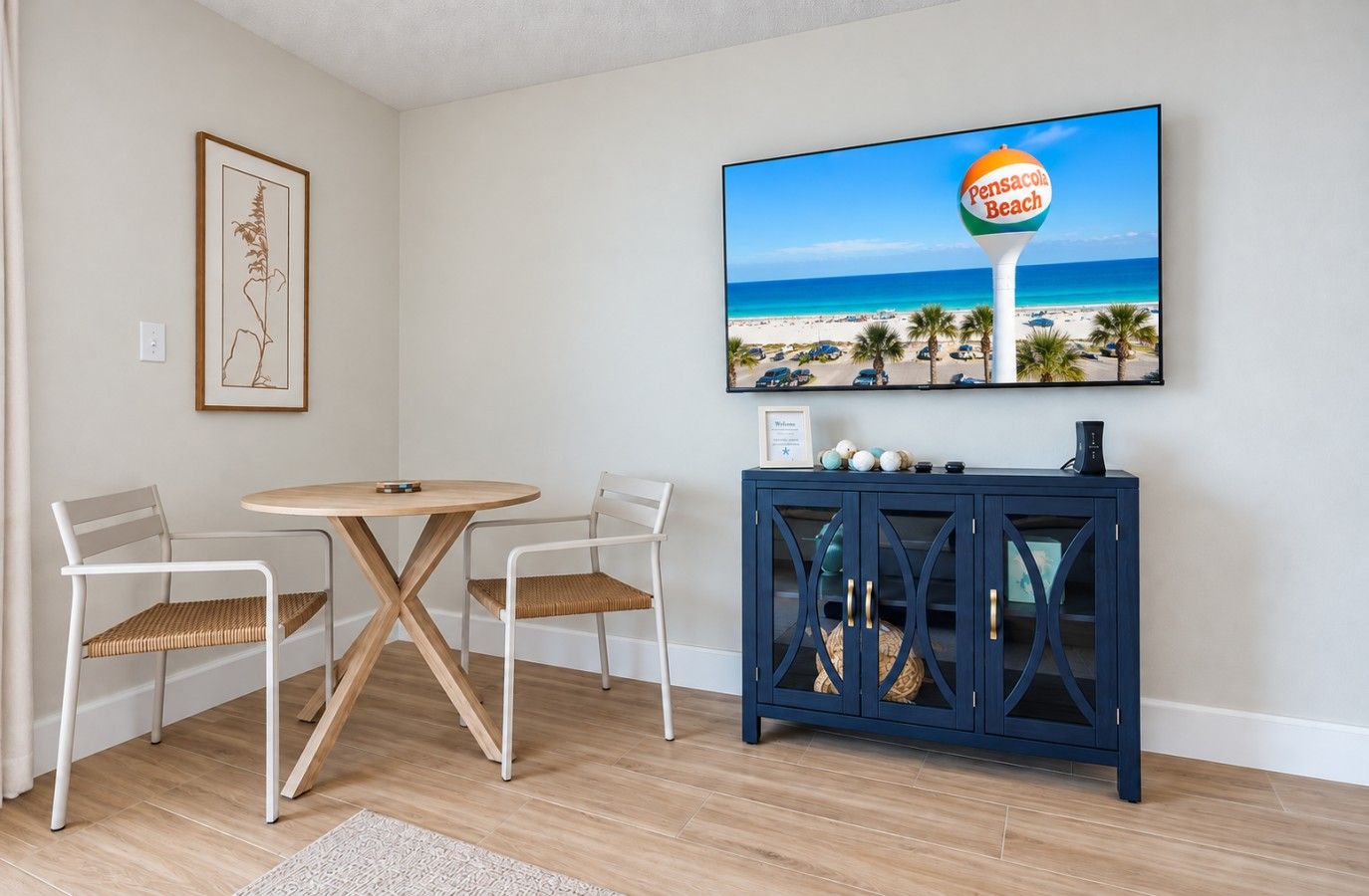 Une pièce comprenant une table ronde en bois et deux chaises, une estampe botanique encadrée accrochée au mur, un buffet bleu foncé aux portes en verre, et un grand écran de télévision diffusant une scène de plage.